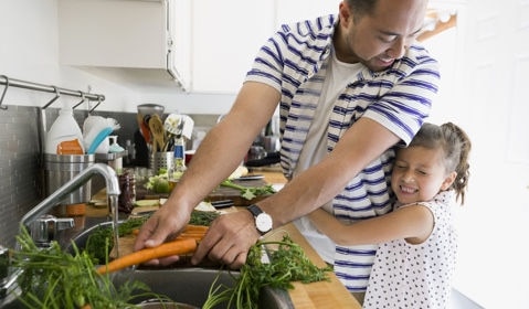 A daughter giving her father a big hug while his washing vegetables.